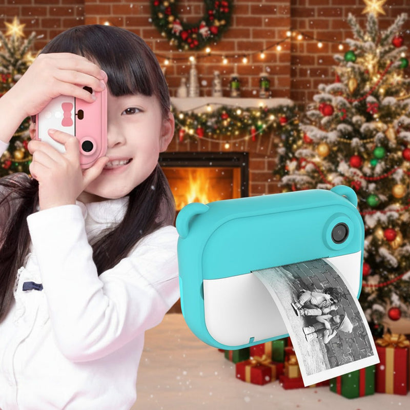 Child using a pink kid instant print camera with a blue instant print camera printing a black-and-white photo in a festive Christmas living room with decorated tree and fireplace